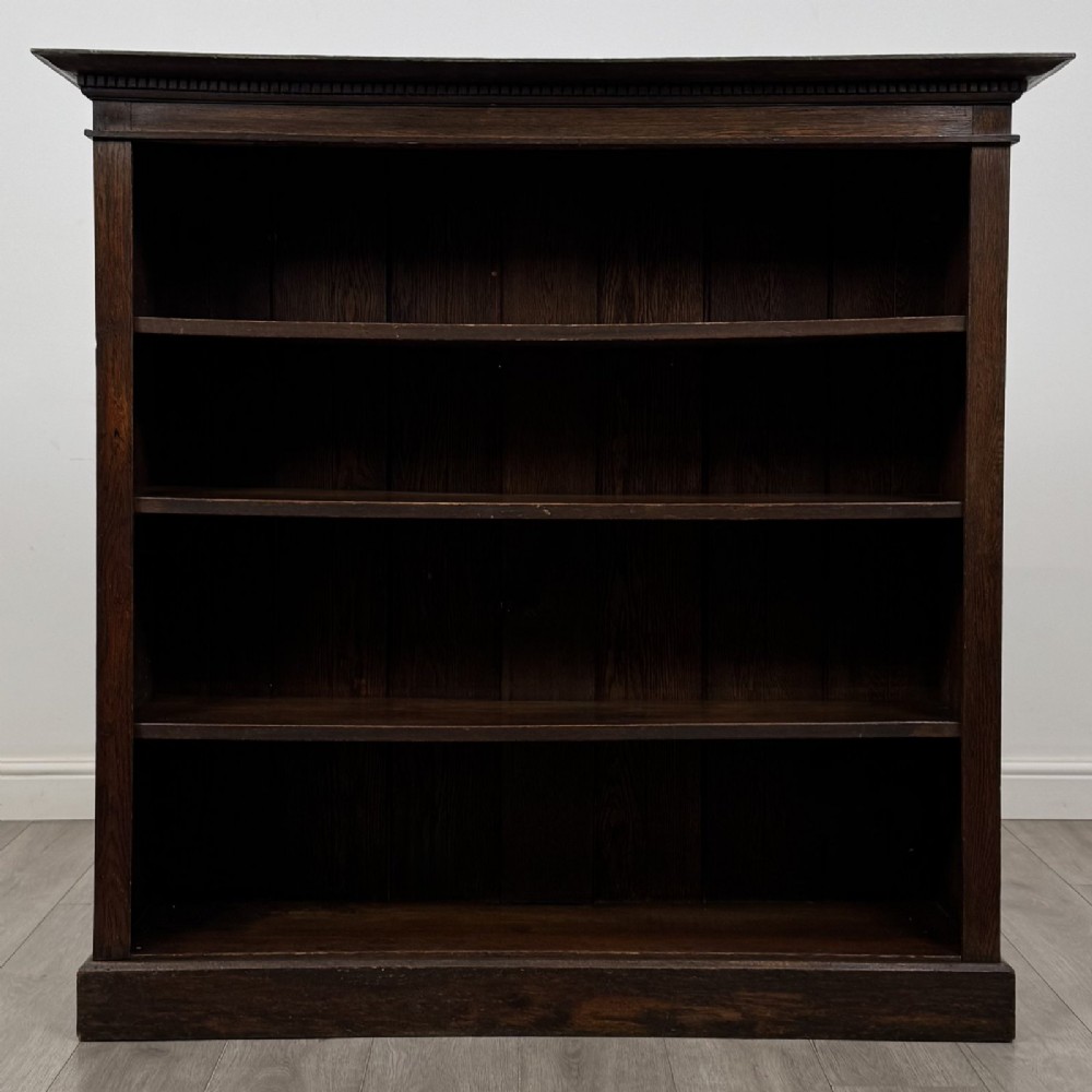 An antique dark oak open bookcase featuring four shelf sections, a dentil carved cornice at the top, and a solid base.
