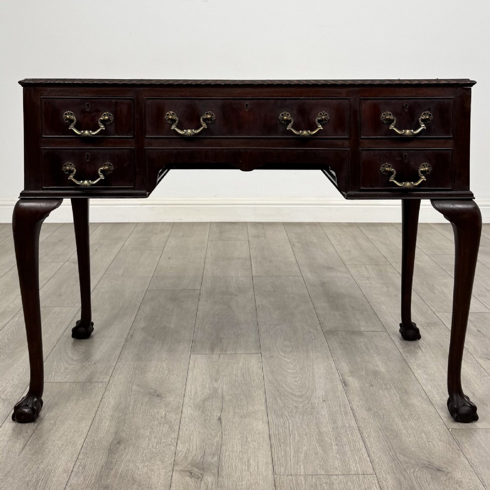An Edwardian dark mahogany writing table with five drawers, decorative brass handles, a carved top edge, and cabriole legs ending in ball and claw feet.