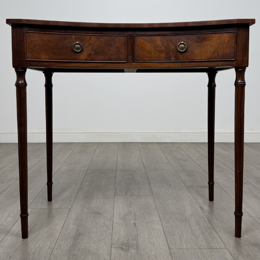 An antique mahogany writing table with a concave bowfront shape, two drawers, brass ring handles, and four reeded legs.