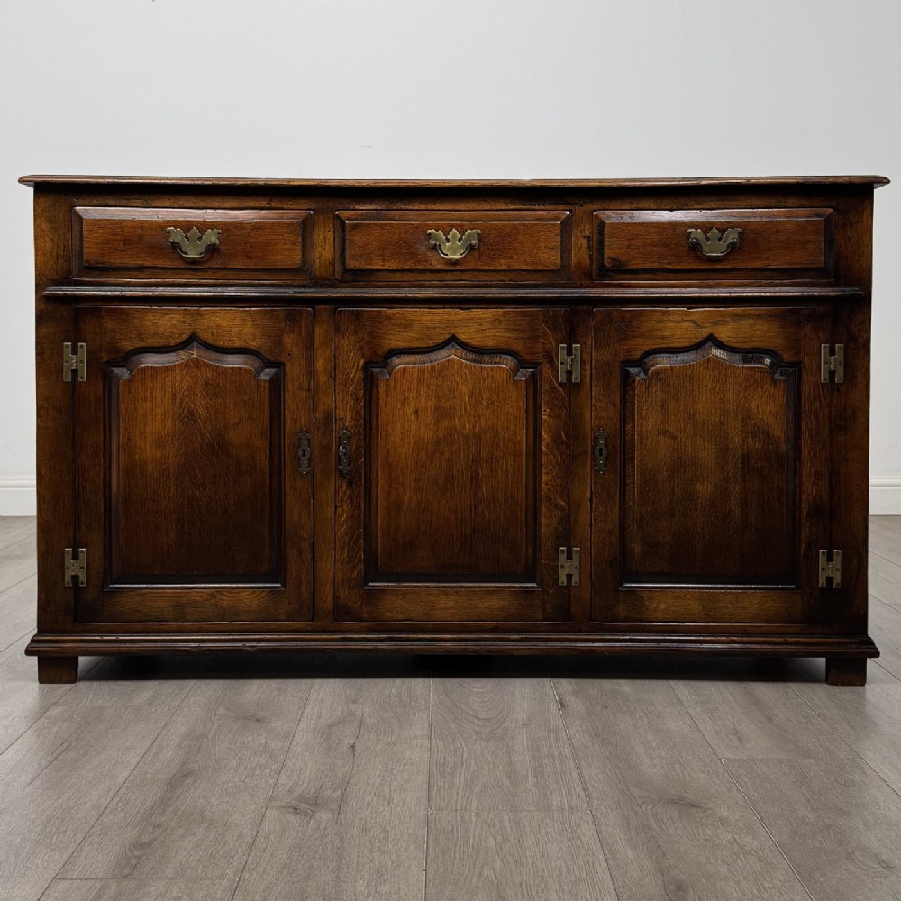 A dark oak sideboard featuring three top drawers with brass handles above three fielded panel cabinet doors with exposed brass H-hinges.