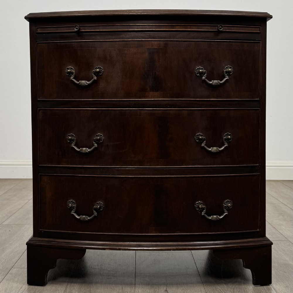 A small dark mahogany bow-front chest of drawers featuring three drawers, a top pull-out slide, and brass swan-neck handles set on bracket feet.