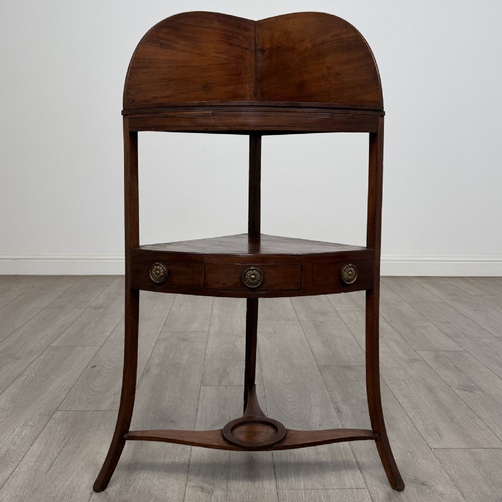A Georgian style mahogany corner washstand with a high curved splashback gallery, central drawer tier with brass rosette handles, and splayed legs joined by a circular lower stretcher.