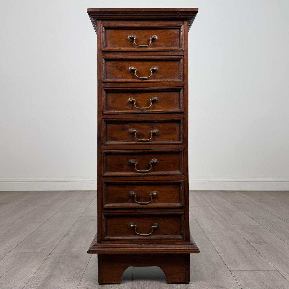 A tall and narrow warm brown mahogany chest of drawers with seven stacked drawers and brass drop handles.