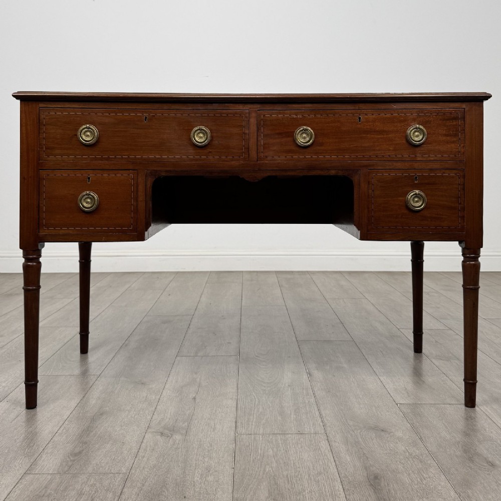 An Edwardian mahogany writing table featuring four drawers with brass ring handles, boxwood stringing inlay, a central kneehole aperture, and four slender turned legs.