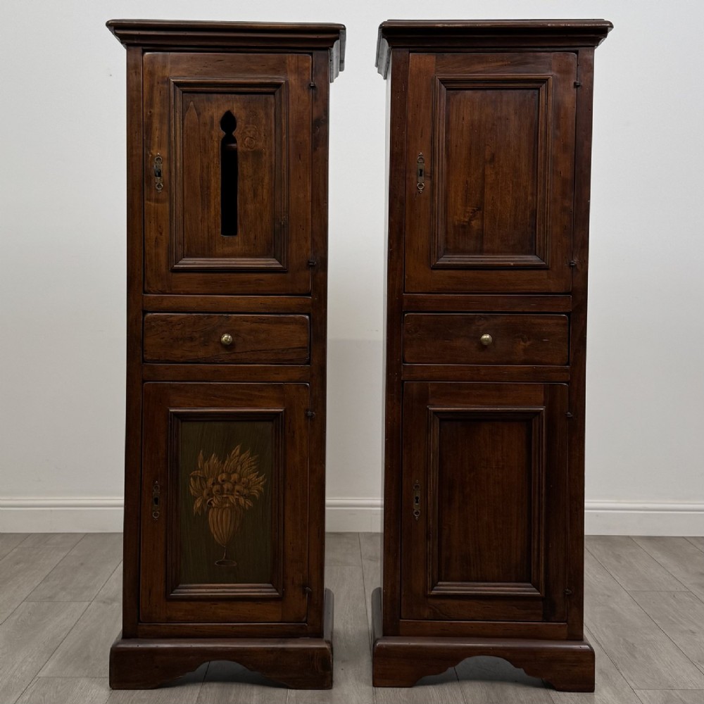 A pair of tall dark brown hardwood cupboards featuring a central drawer and panelled doors, with one lower panel decorated with a painted urn motif.