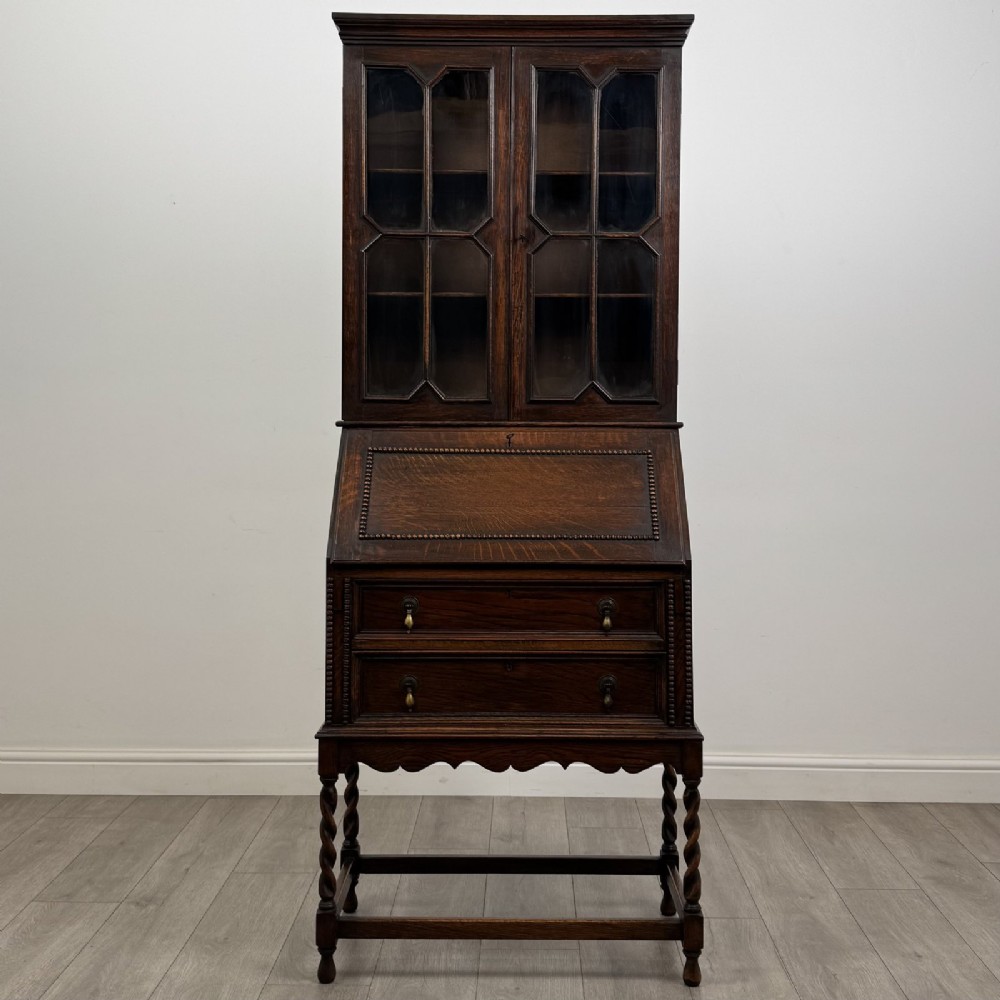 A dark oak Jacobean style bureau bookcase featuring twin glazed cabinet doors above a slant-front writing desk with two lower drawers and barley twist legs.