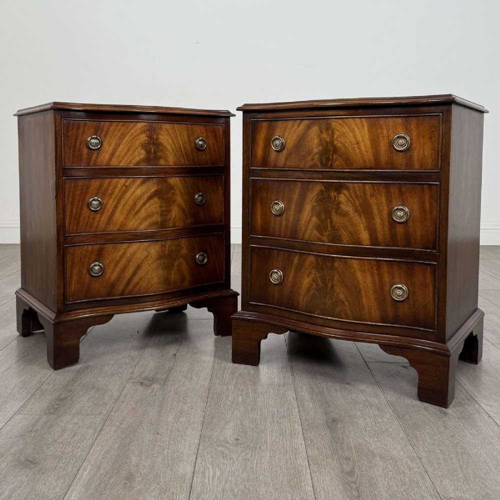 A matching pair of Georgian style mahogany bedside chests with serpentine fronts, three drawers each, brass ring handles, and bracket feet.