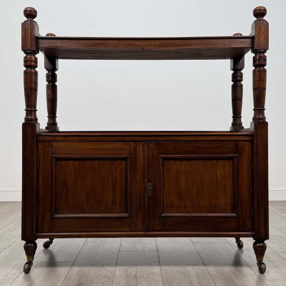 A Victorian mahogany buffet trolley featuring an upper open shelf supported by turned column pillars over a lower two-door panelled cupboard set on brass castors.