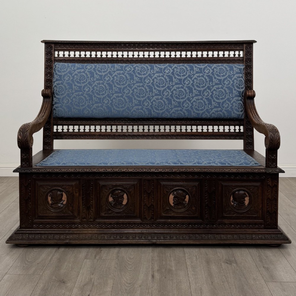 A dark carved oak hall bench with patterned blue upholstered seat and backrest cushions, spindled galleries, and relief-carved profile medallions across the base.