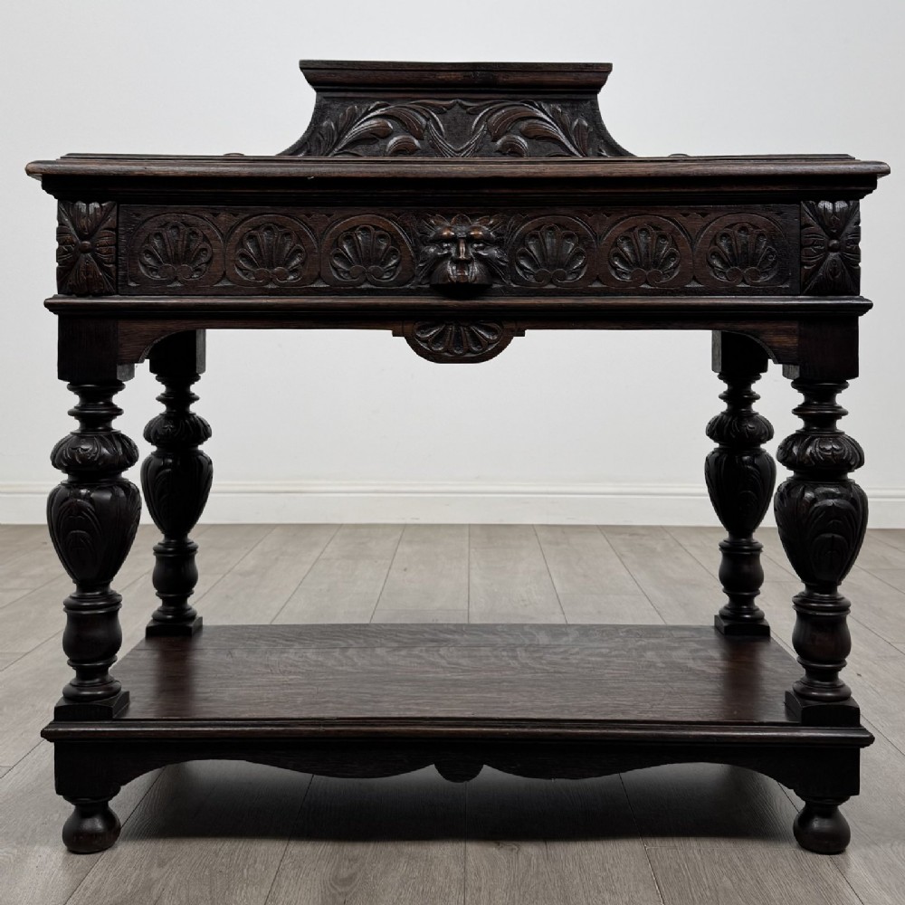 A dark carved oak side table featuring a Green Man mask on the front frieze, turned legs, a low undertier, and a raised carved gallery back.