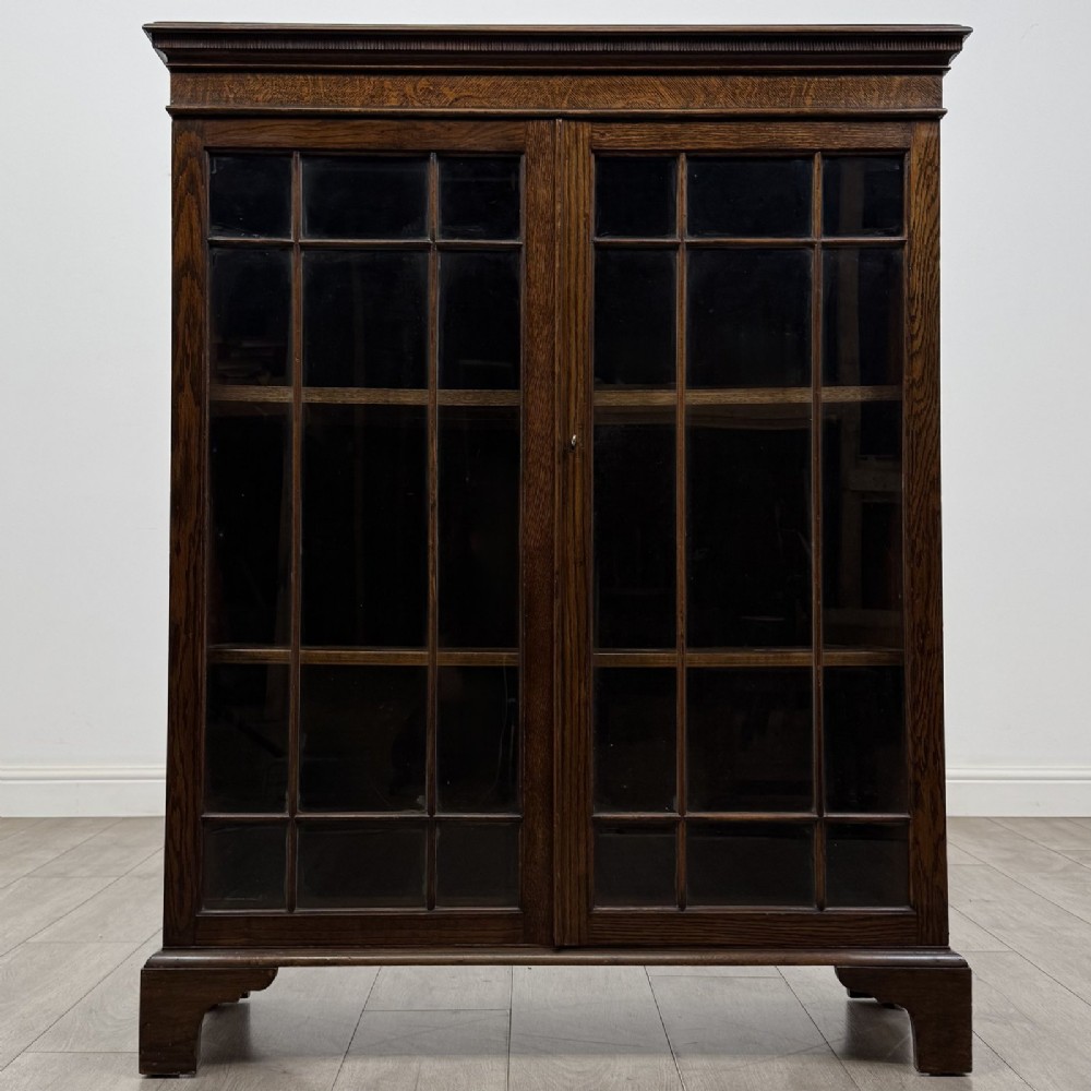 A dark oak Georgian style display cabinet featuring twin glazed doors with rectangular timber mullions and bracket feet.