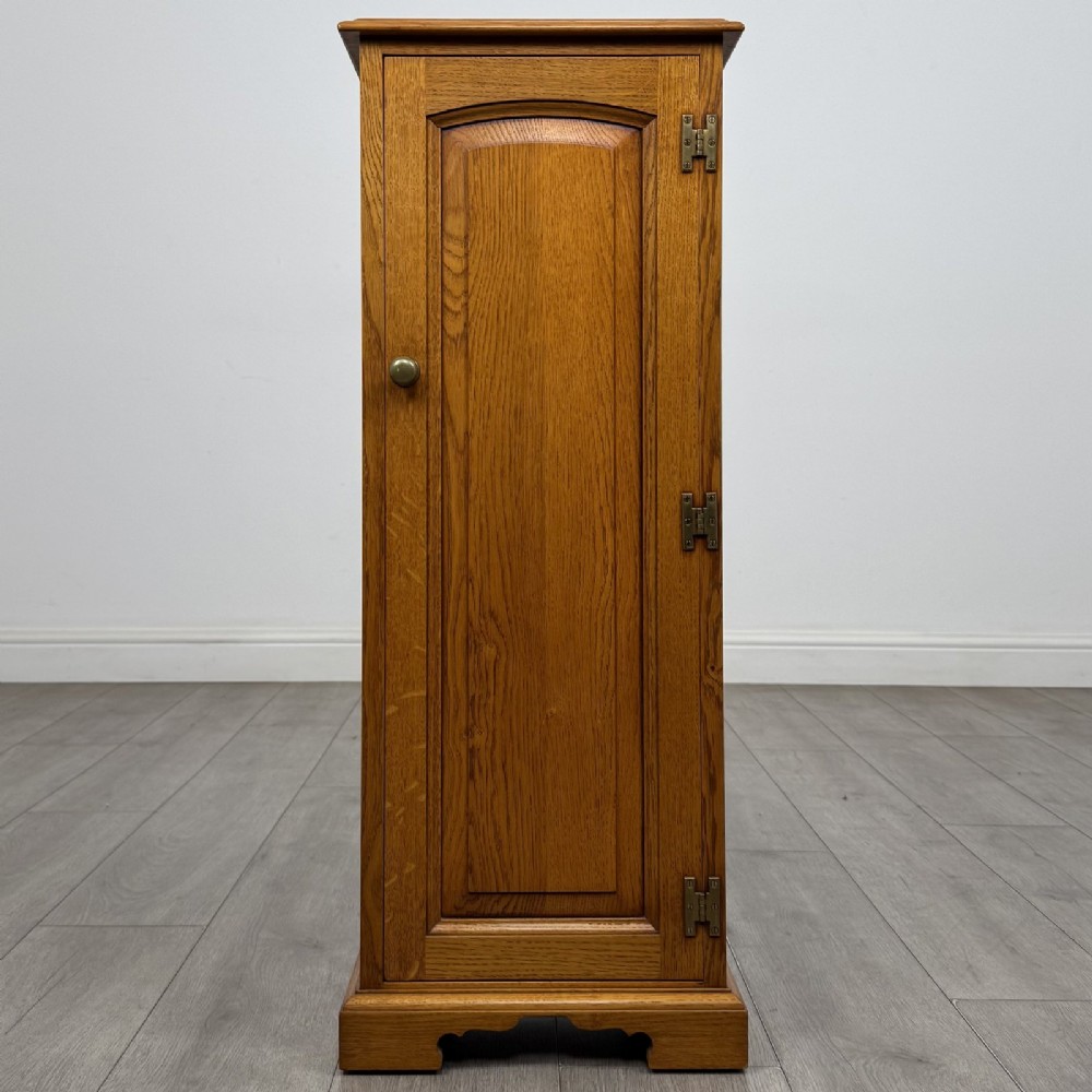 A tall, narrow warm oak cupboard featuring a single arched fielded panel door with three brass H-hinges and a small brass door knob.