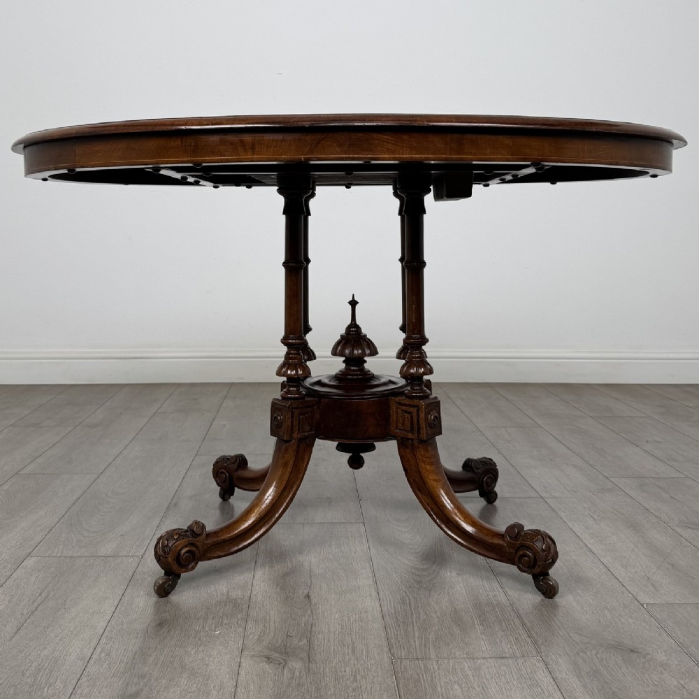 A Victorian burr walnut tilt-top loo table featuring an oval top supported by four turned columns and carved quadform legs with scroll feet.