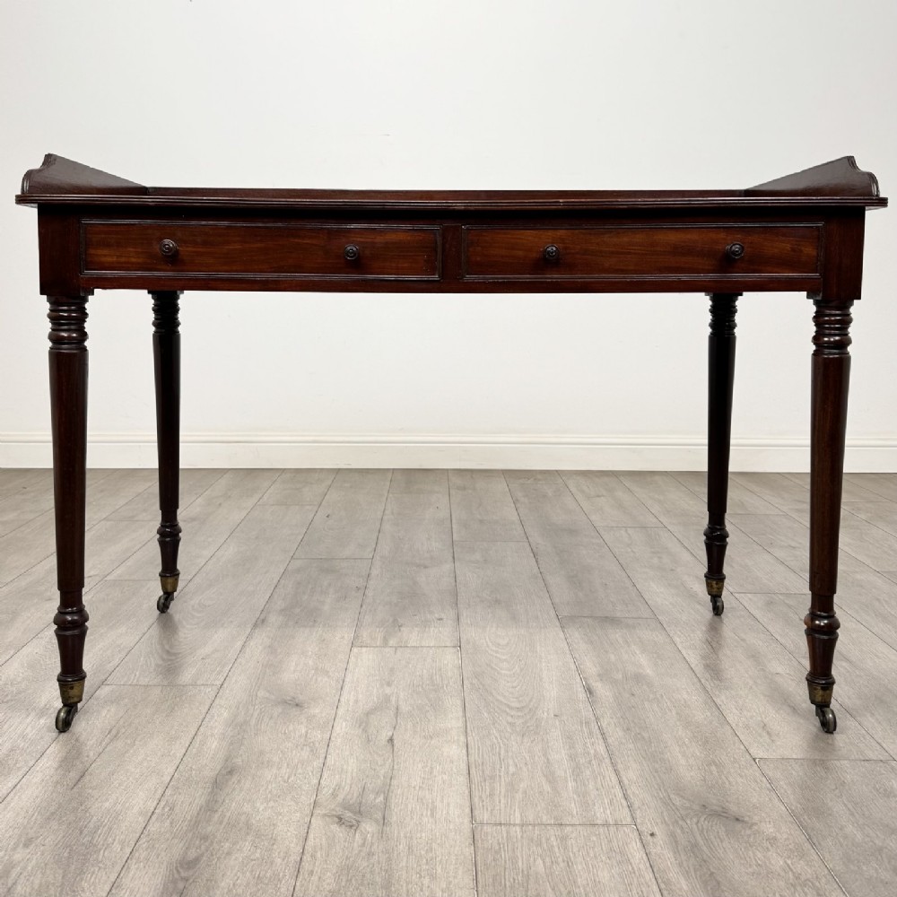 A dark mahogany Georgian writing desk featuring two frieze drawers with wooden knobs, turned legs on brass castors, and a raised gallery edge.