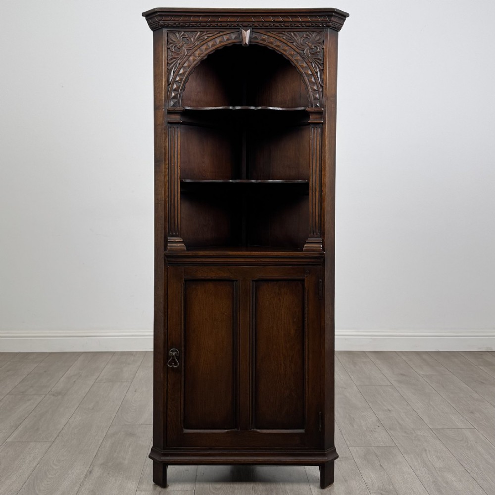 A tall dark oak Jacobean style corner cabinet featuring an upper open section with an arched alcove and two shelves above a single panelled cupboard door.