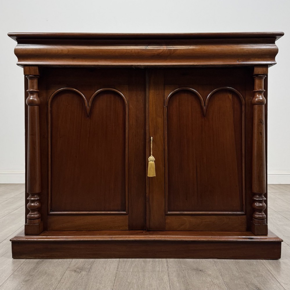 An antique Victorian dark mahogany side cabinet featuring double arched panel doors, flanking turned columns, a plinth base, and a concealed frieze drawer.