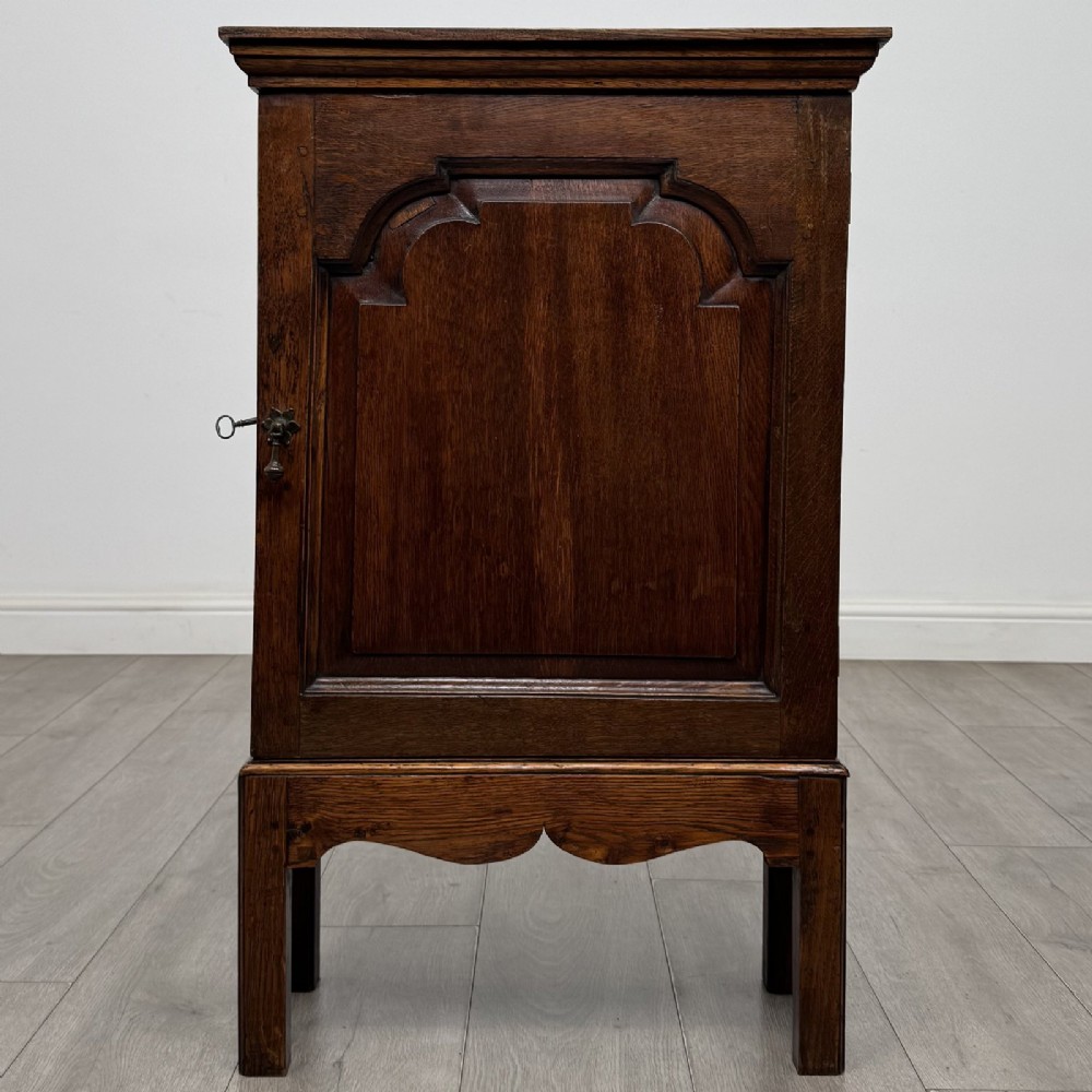 A small dark Georgian oak spice cupboard on a stand with an arched fielded panel door and brass keyhole.