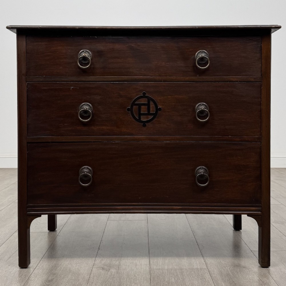 A dark mahogany Arts and Crafts chest of drawers with three drawers, circular brass ring handles, and a central circular motif.