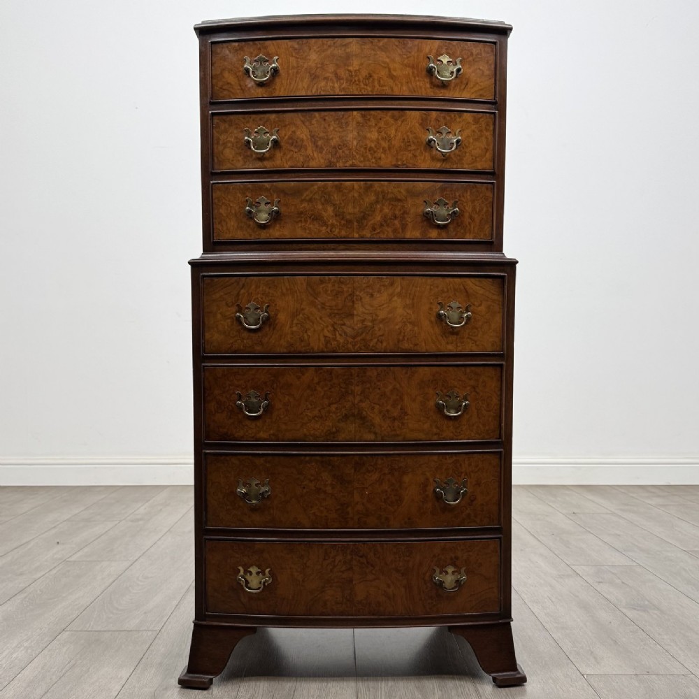 A Georgian style burr walnut tallboy featuring a bow-front silhouette, seven stacked drawers with brass handles, and bracket feet.