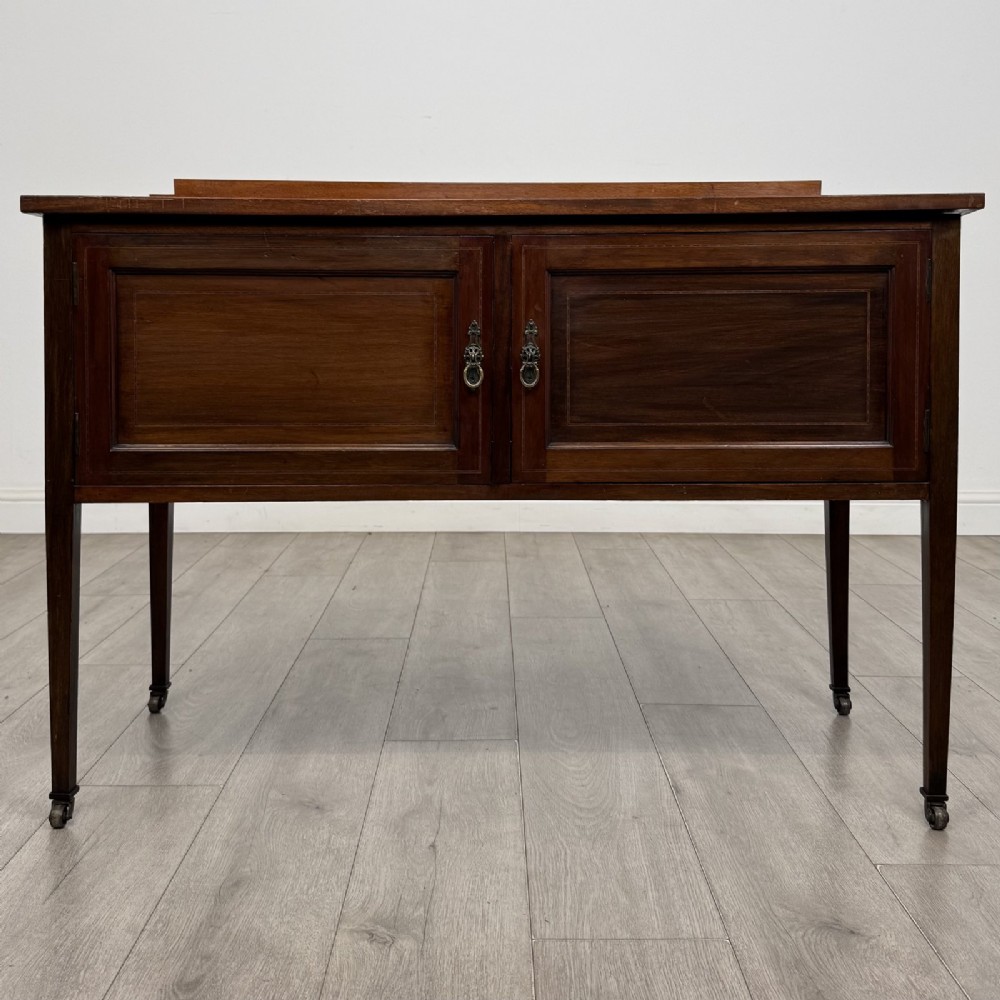 An Edwardian mahogany side cabinet featuring double panelled doors with line inlay, square tapered legs, brass drop handles, and brass castors.