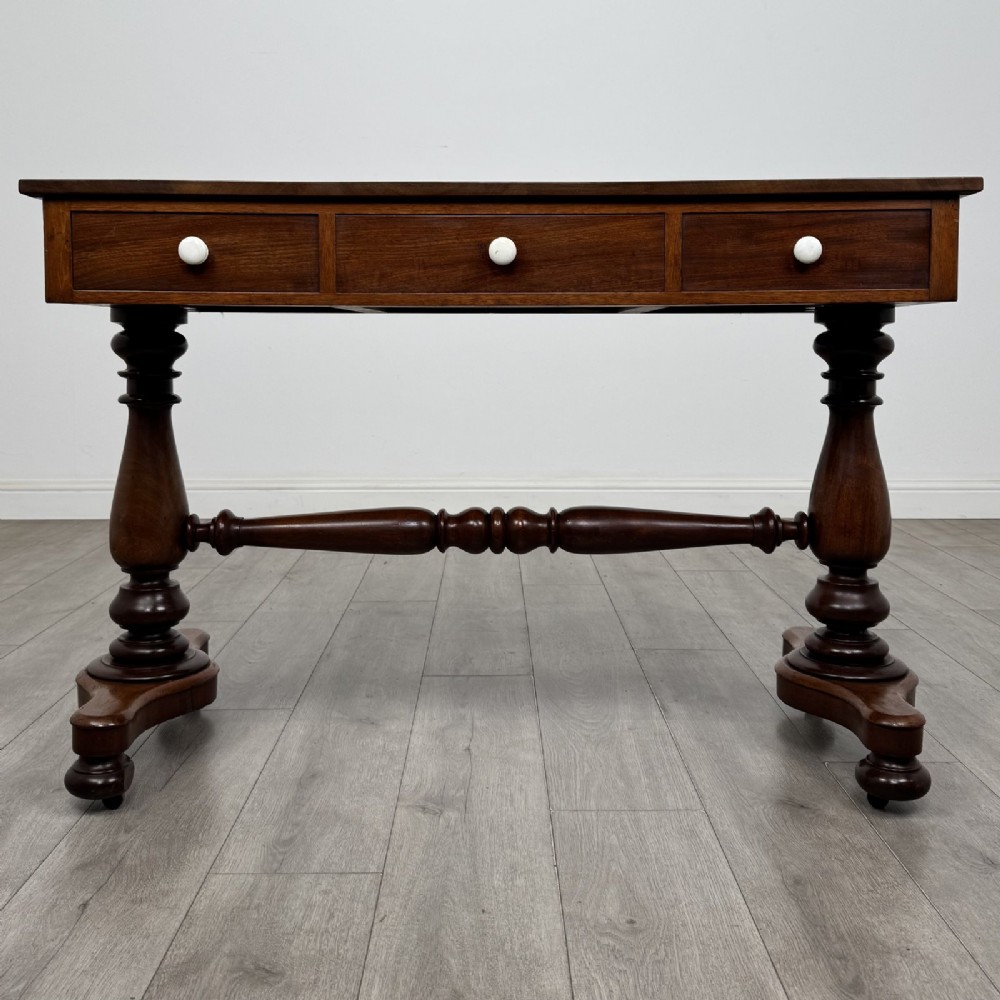 A Victorian mahogany writing table featuring three front drawers with white porcelain knobs, set on turned baluster legs with a central stretcher bar and castors.