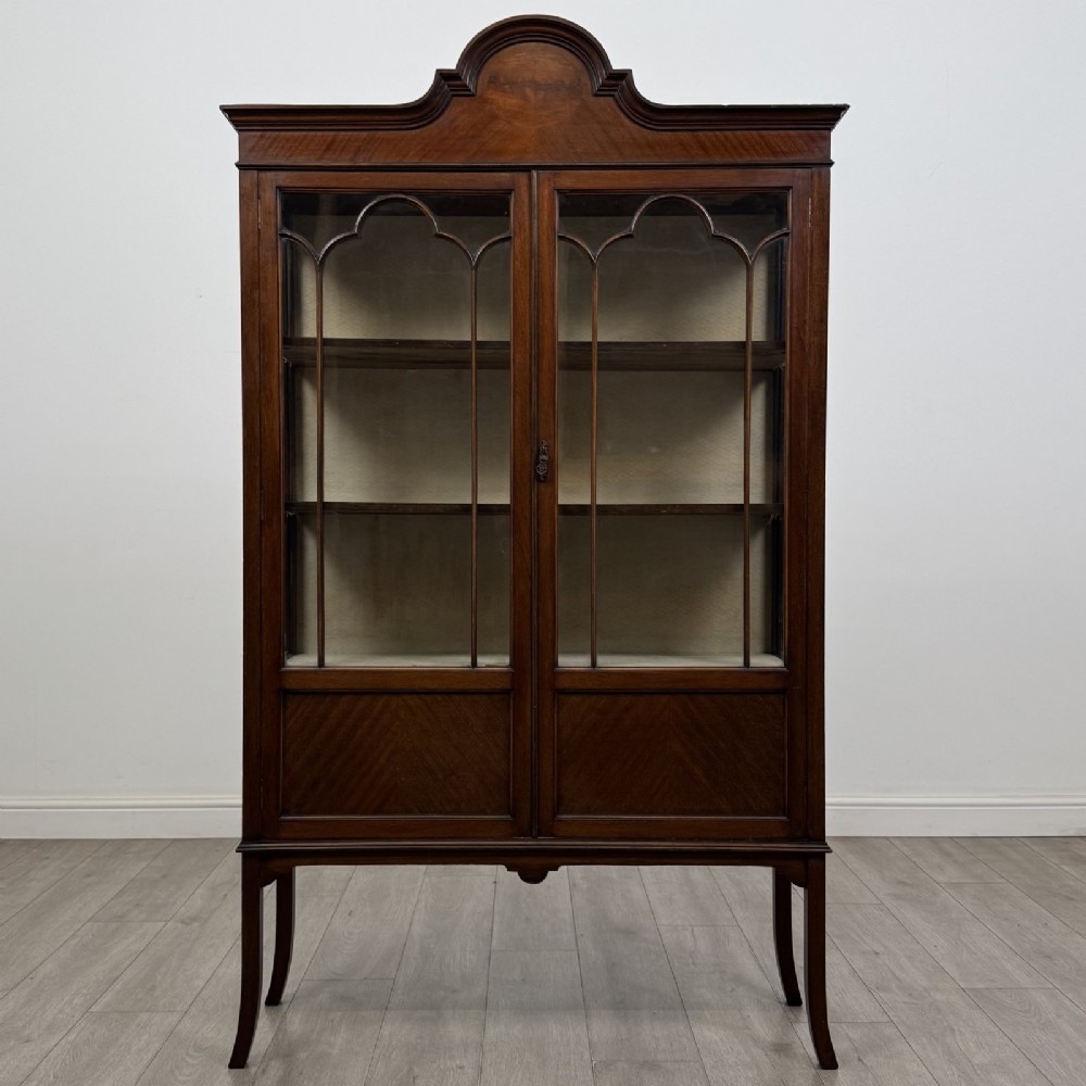 An Edwardian mahogany display cabinet featuring two astragal glazed doors, a shaped arched cresting, and lower veneered panels supported on slender splayed legs.