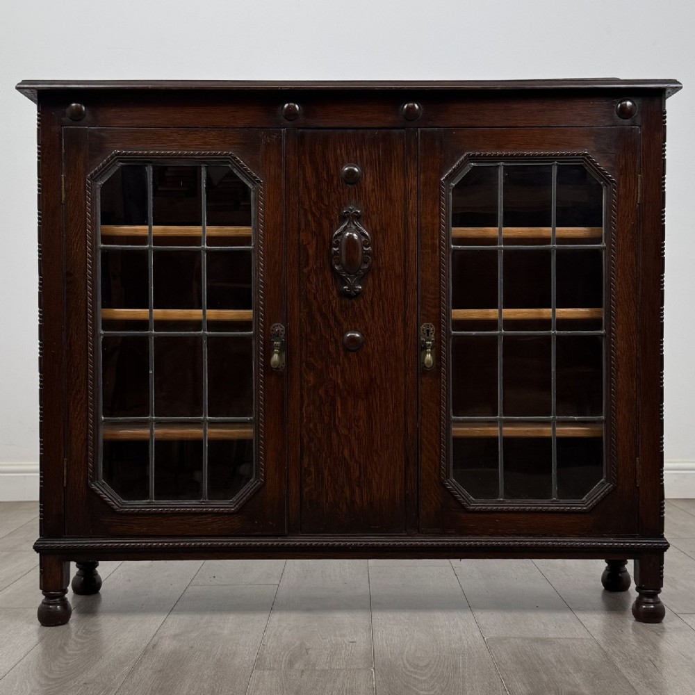 A dark oak Jacobean style display cabinet featuring twin leaded glass doors, central carved bosses, internal shelving, and turned feet.