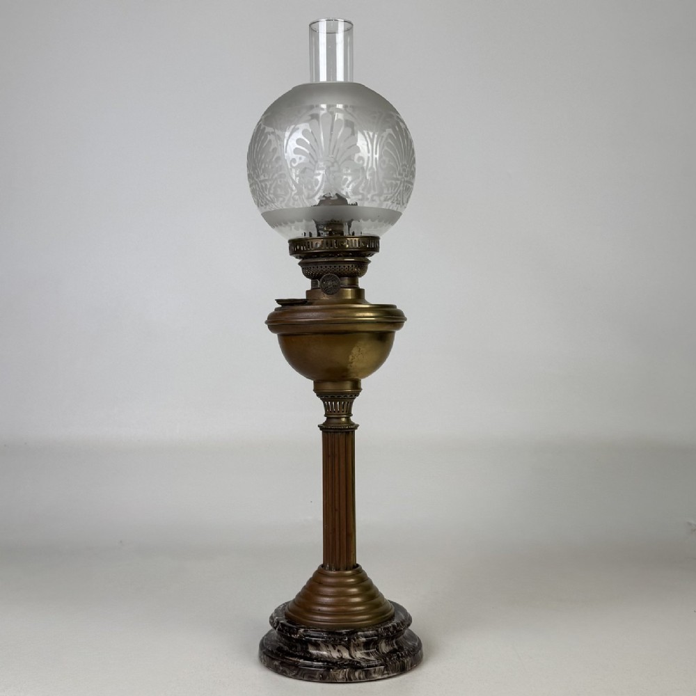 An antique brass column oil lamp featuring an etched glass globe shade, clear chimney, brass reservoir, fluted stem, and dark veined marble base.