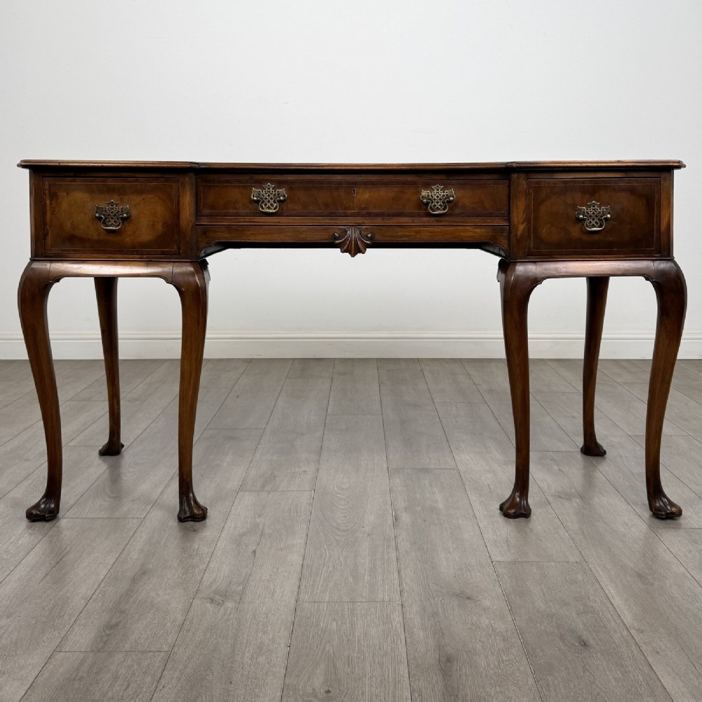 A Queen Anne style walnut writing table featuring three drawers, decorative brass handles, carved cabriole legs, and a central shell motif on the lower apron.