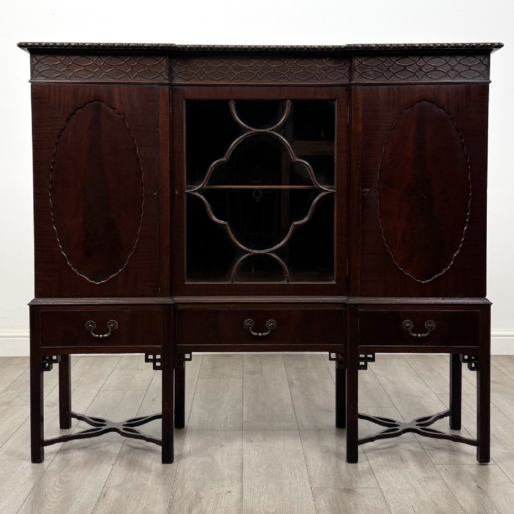 An Edwardian dark mahogany breakfront cabinet featuring a central glazed door with geometric glazing bars flanked by two solid oval panel doors over three drawers and cross-stretched legs.