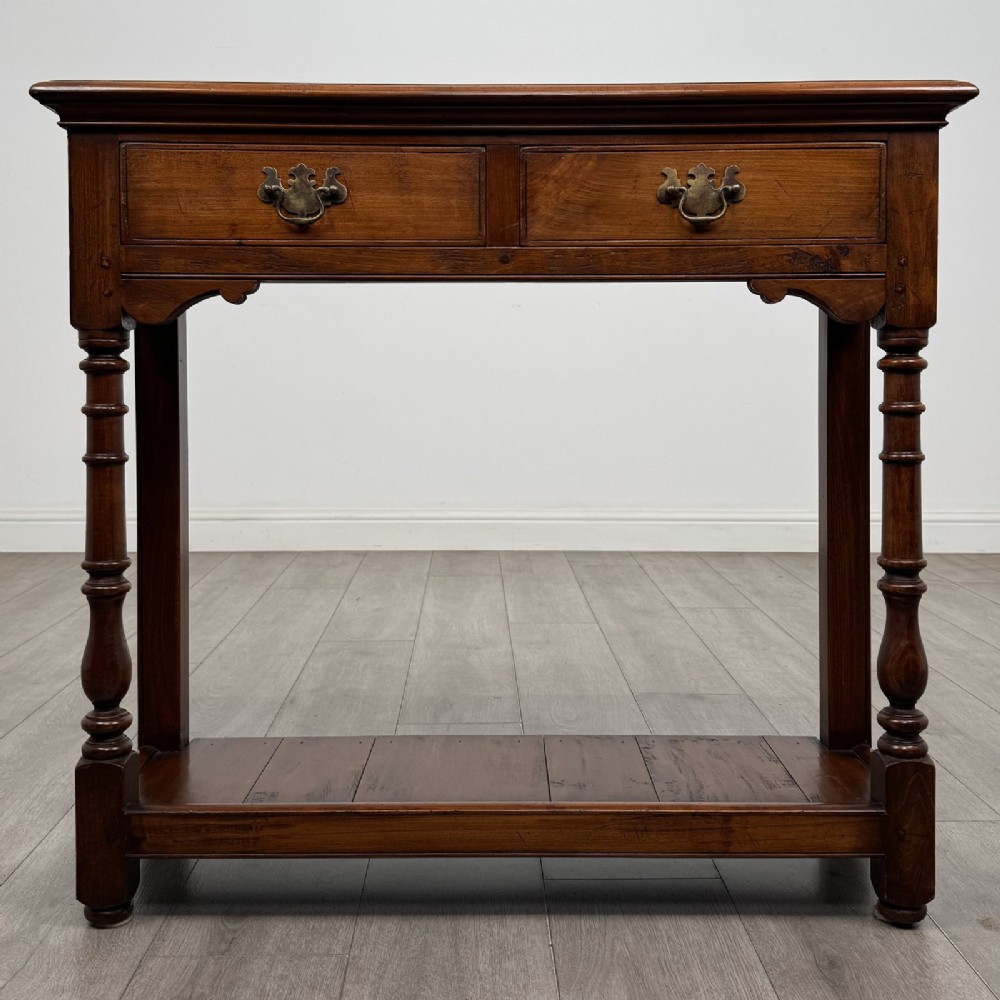 A narrow dark wood console table featuring two drawers with brass handles, turned front legs, and a lower potboard shelf.