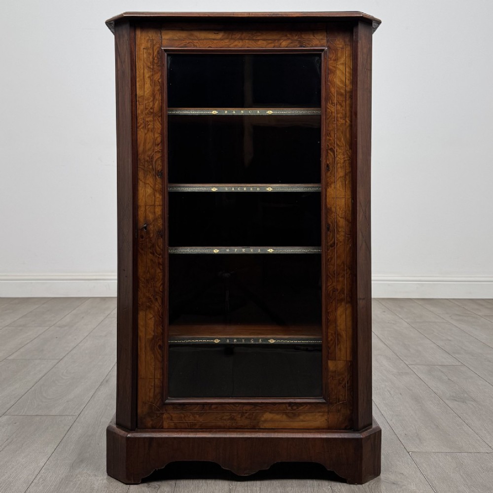 A Victorian burr walnut music cabinet featuring a single glazed door revealing internal shelves with gilt-embossed text labels.