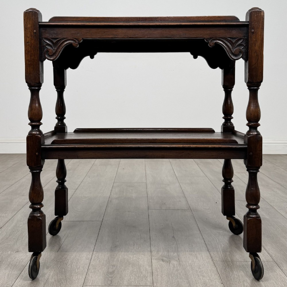 A dark brown oak two-tier tea trolley featuring turned supports, carved corner spandrels, and metal castors on the feet.