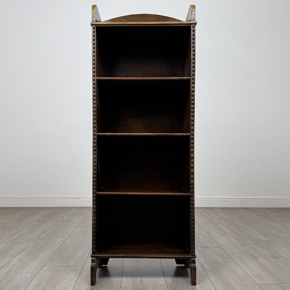 A tall, narrow dark oak open bookcase featuring four fixed shelves, beaded front stiles, and a low arched top gallery rail.
