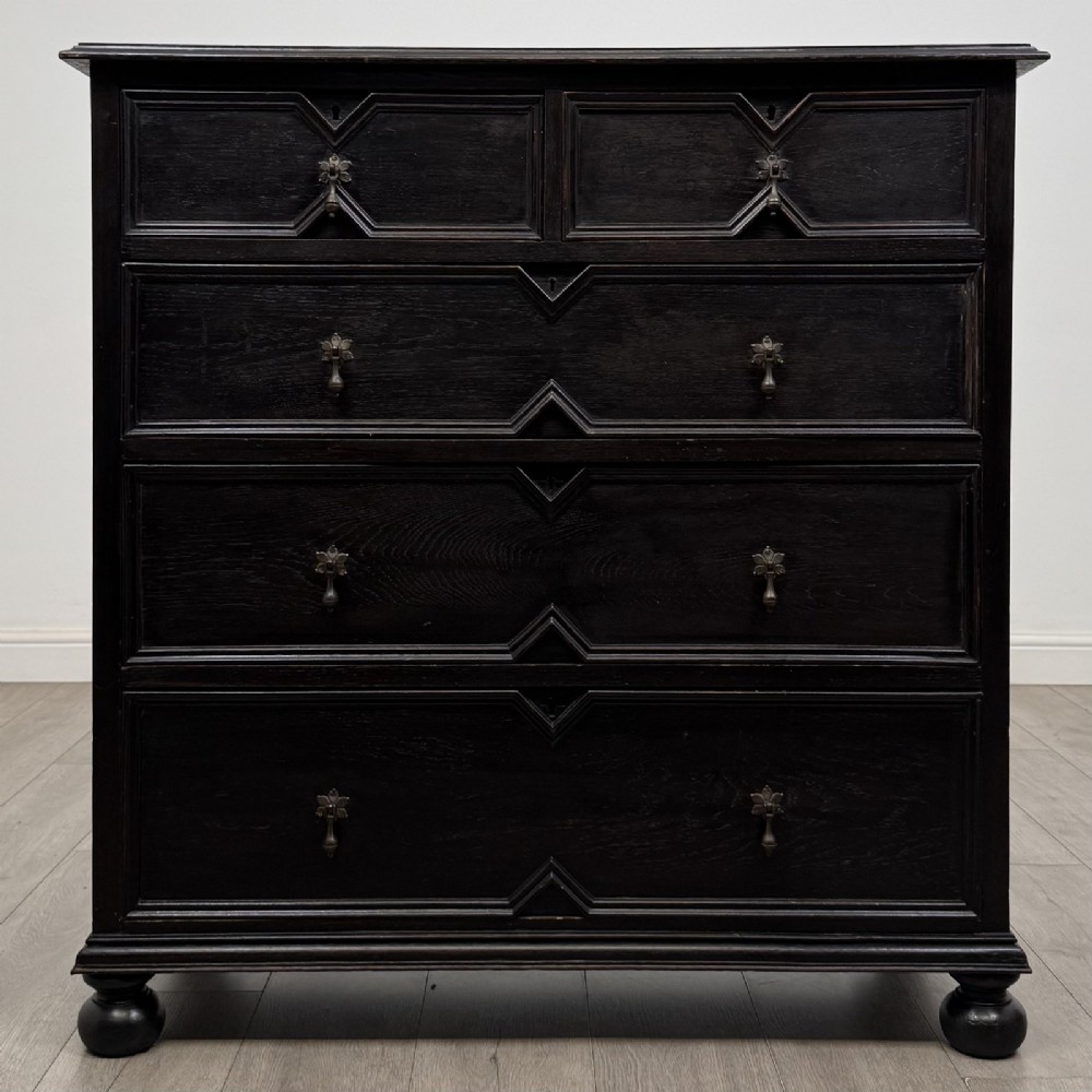 An ebonised oak chest of drawers featuring geometric moulded panel fronts, brass drop handles, and turned bun feet.