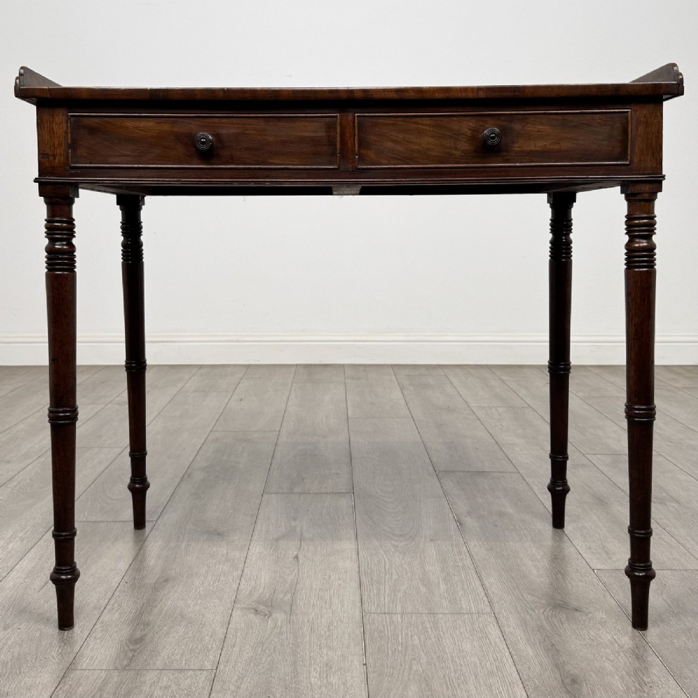 An antique dark mahogany Regency writing table featuring two front drawers with turned wooden knobs and four tapered ring-turned legs.