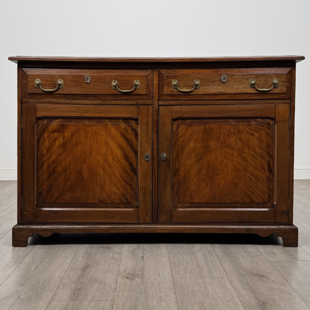 A Georgian style warm brown oak dresser base featuring two drawers with brass drop handles above two panelled cupboard doors, raised on bracket feet.