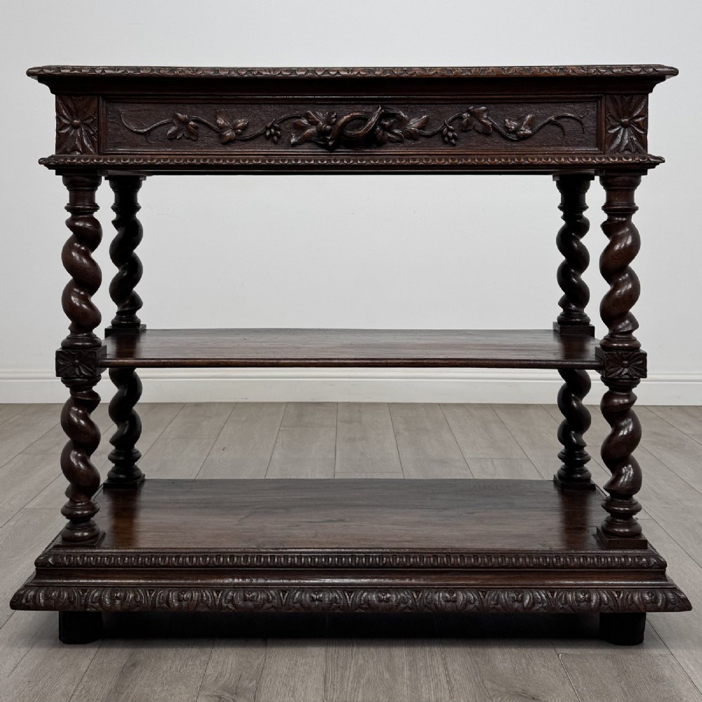 A dark oak three-tier buffet featuring relief foliage carving across the top drawer frieze and four barley twist corner support legs.