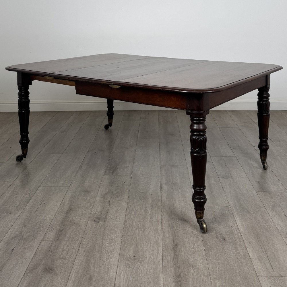 An antique William IV mahogany extending dining table featuring a dark timber top with rounded corners, turned legs, and brass castors.