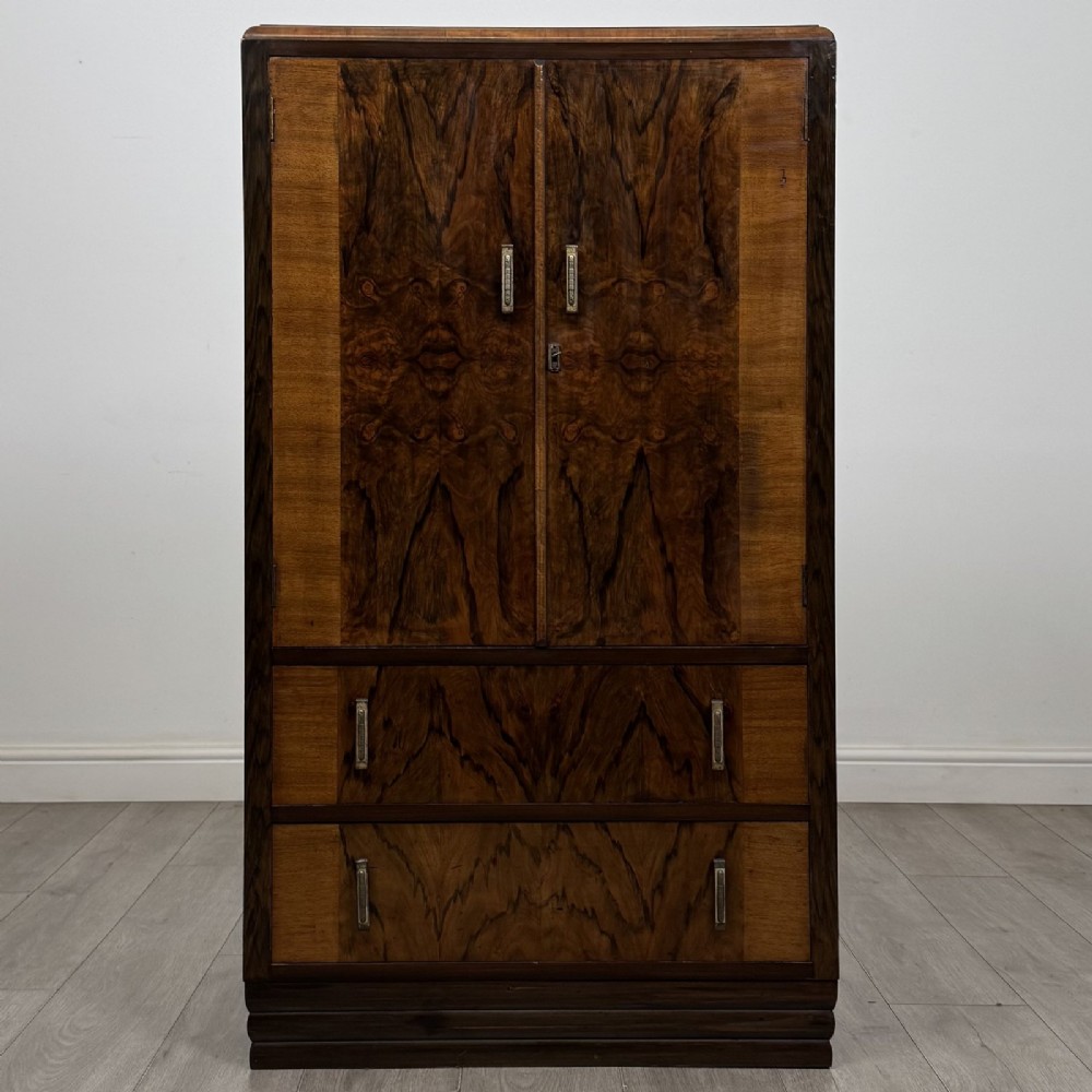 An Art Deco walnut tallboy cabinet featuring highly figured book-matched veneer, double doors over two long drawers, vertical metal handles, and a stepped plinth base.