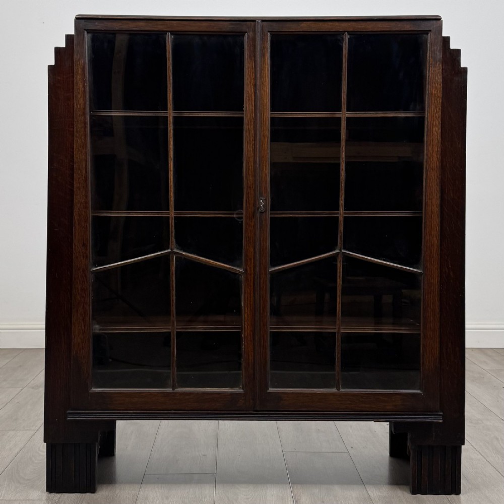A dark oak Art Deco display cabinet with two glazed doors, geometric glazing bars, stepped side shoulders, and reeded square feet.