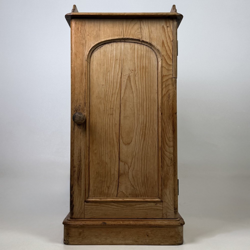 An antique natural pine pot cupboard featuring a single arched panel door, turned wooden knob handle, side top galleries, and a solid plinth base.