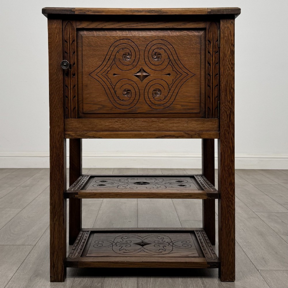 A dark carved oak cabinet featuring a single door adorned with scrolling strapwork designs situated above two open lower shelves.