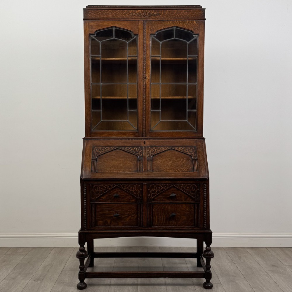 A dark Jacobean style oak bureau bookcase with twin leaded glass upper doors, carved fall-front desk, two lower drawers, and turned legs.
