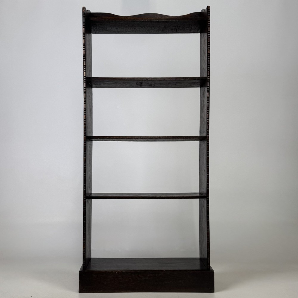 A tall and narrow early 20th-century dark oak open bookcase with five shelf levels and a shaped top gallery.