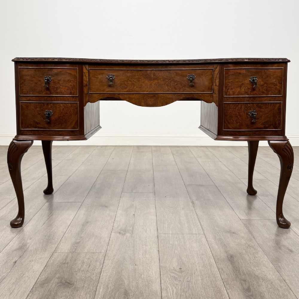 A Queen Anne style burr walnut writing desk featuring a serpentine front, five drawers with brass handles, and carved cabriole legs.