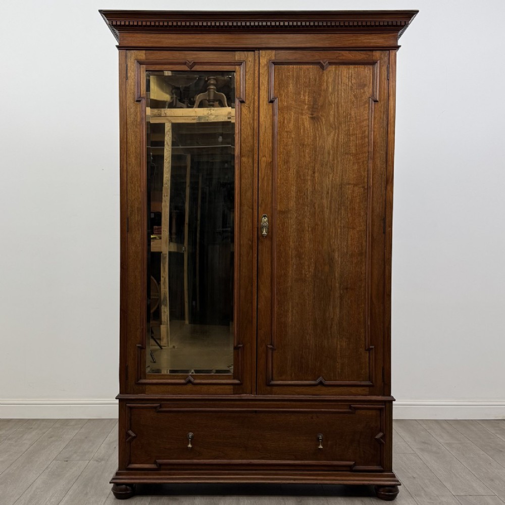 An antique early 20th-century walnut double wardrobe featuring a full-length mirror door, panelled right door, geometric mouldings, and a single lower drawer.