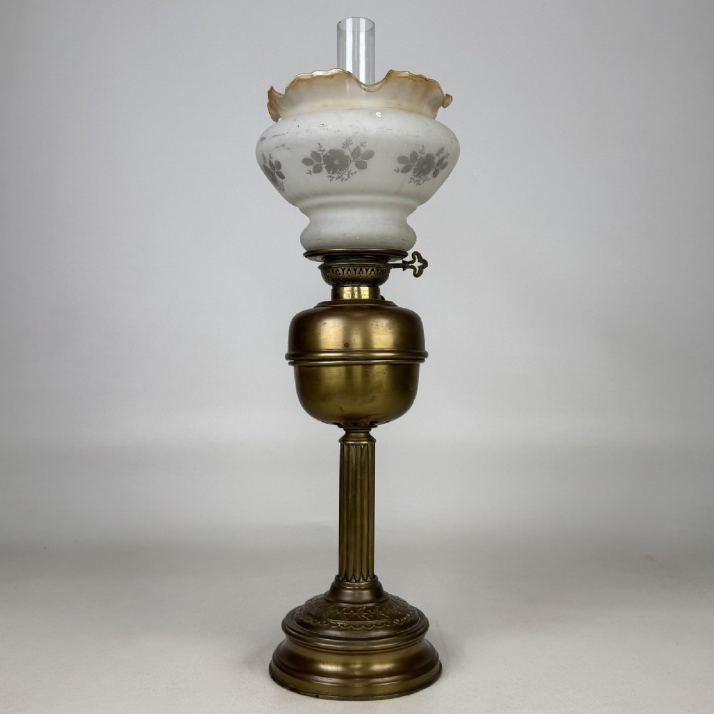 An antique brass oil lamp featuring a reeded pedestal column, a clear glass chimney, and a frilled frosted glass shade decorated with floral etching.