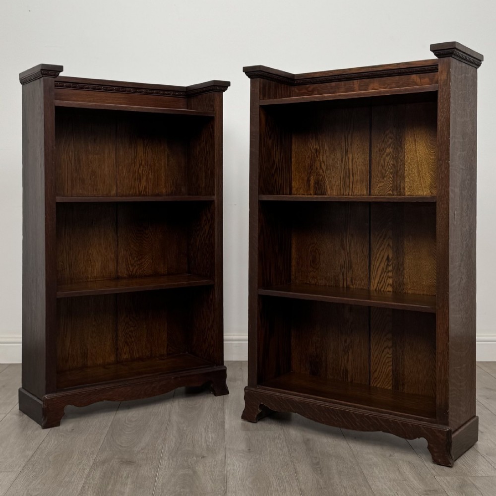 A matching pair of dark oak Edwardian open bookcases featuring carved cornices, three shelves, and shaped plinth bases.