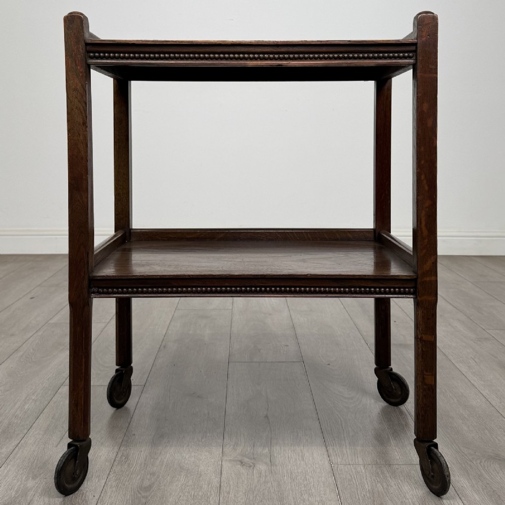 A dark oak two-tier tea trolley featuring beaded moulding along the edges and small dark castors on four straight legs.